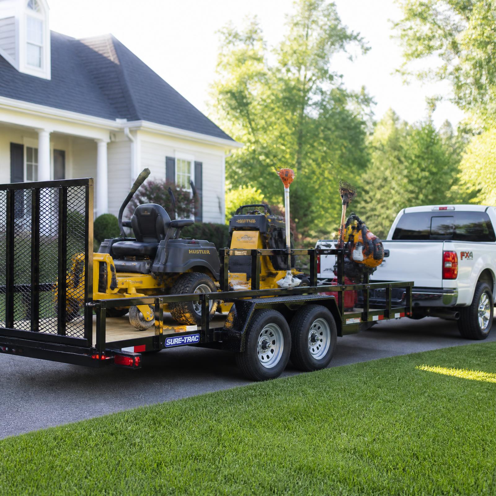 A professional landscaping utility trailer loaded with mowers and equipment, hitched to a white pickup truck on a residential lawn